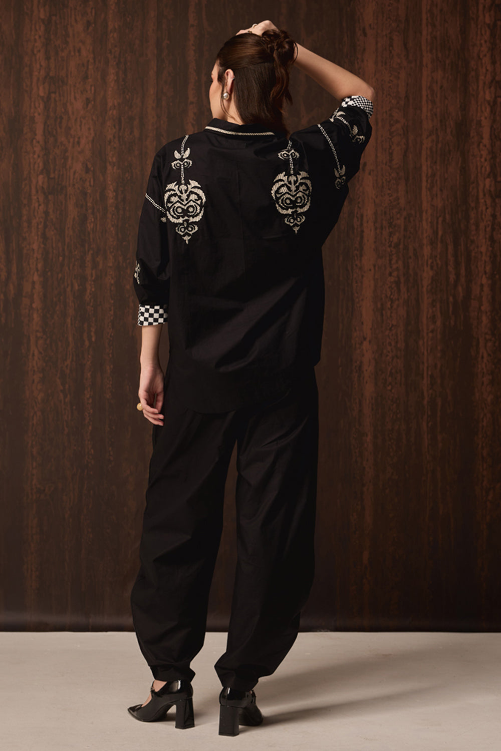 A model is shown from behind wearing a black long-sleeve top and matching wide-leg pants, featuring ornate white embroidery on the back and checkered cuffs, set against a dark wood-paneled background.