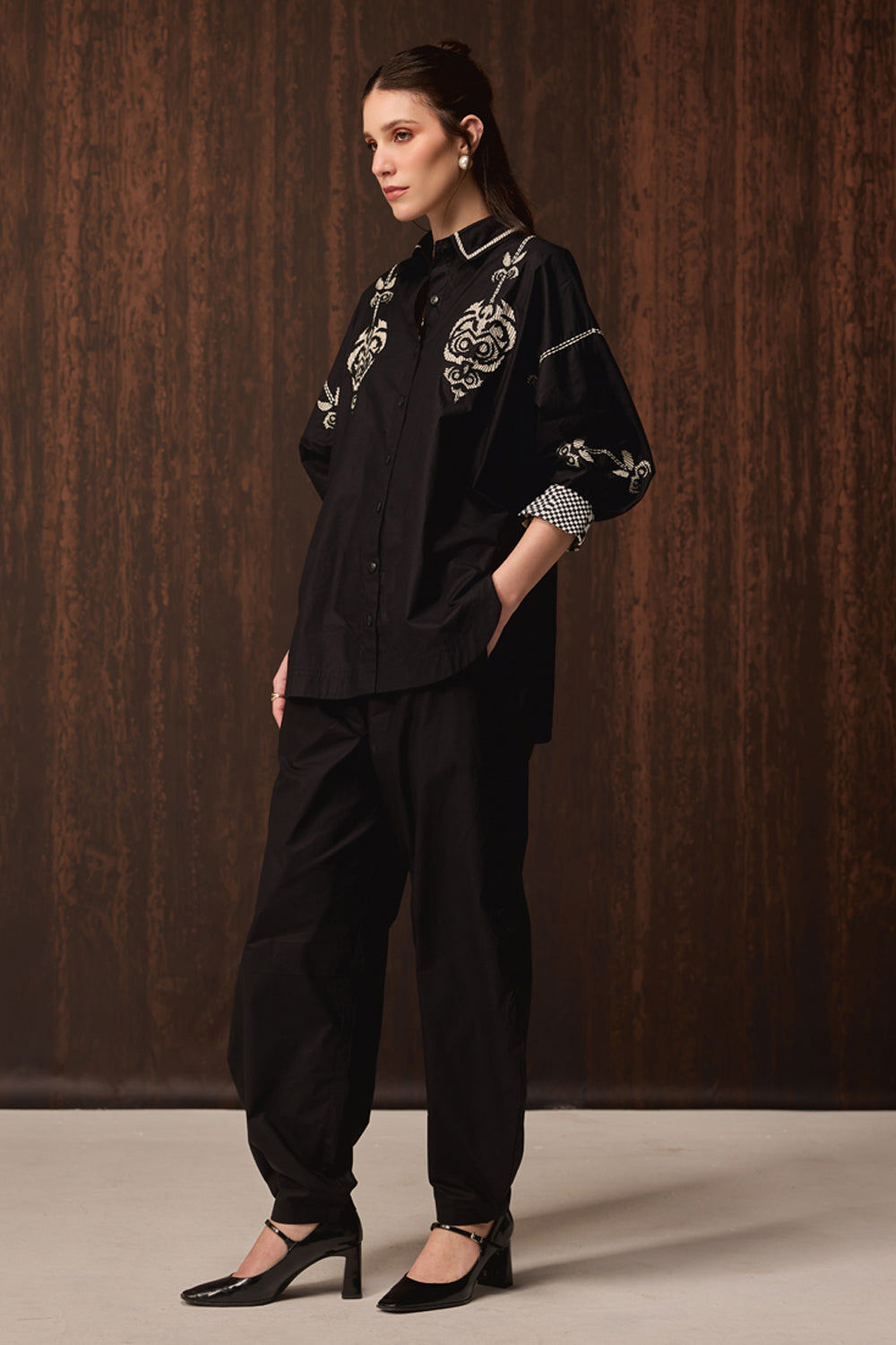 A woman models a black embroidered button-up shirt and matching black trousers against a dark wood-paneled studio background.