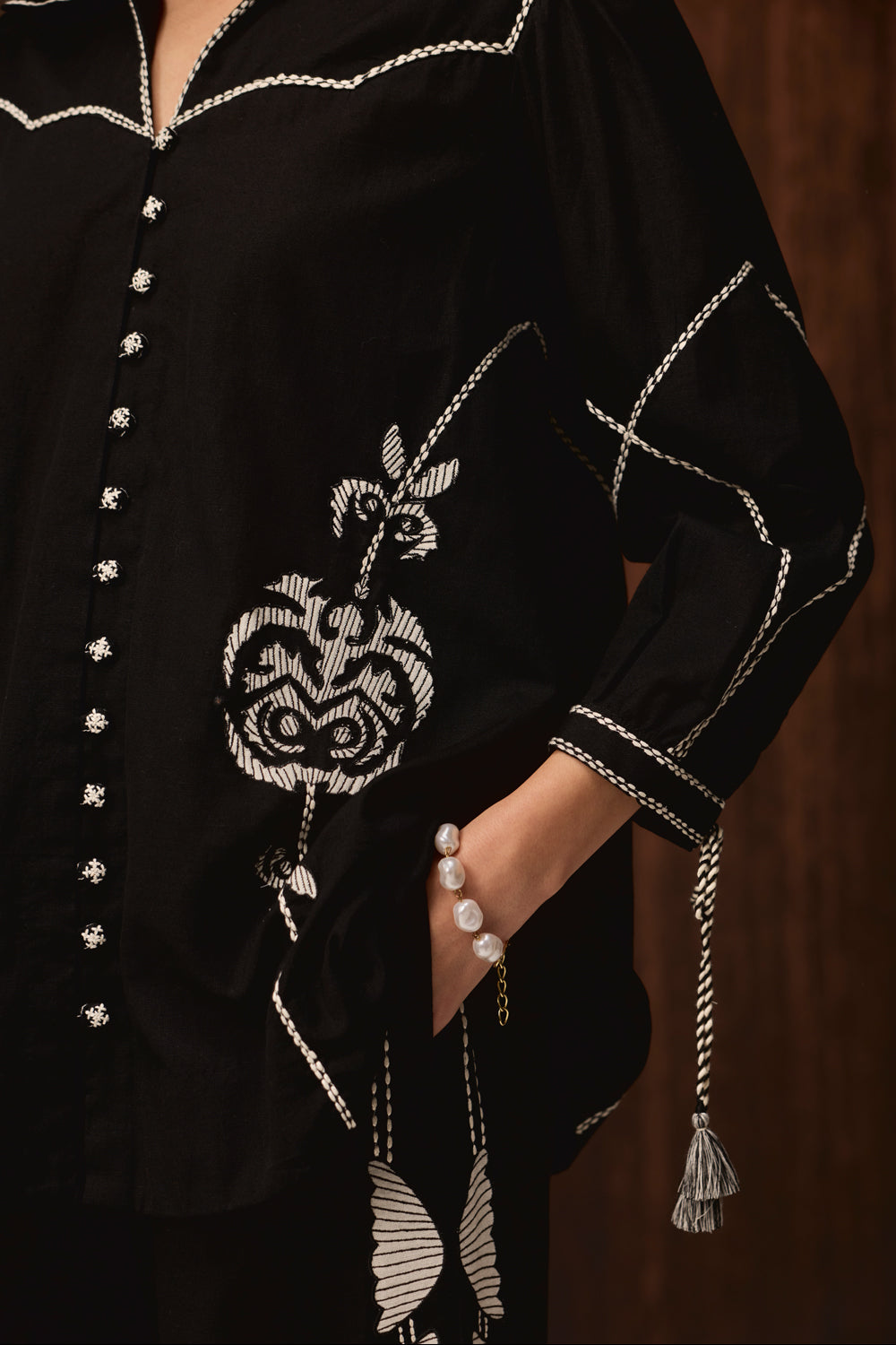 A close-up of a black embroidered tunic or blouse with white and silver thread detailing, featuring a decorative motif on the front, a tassel tie at the sleeve cuff, and a hand tucked into a pocket.