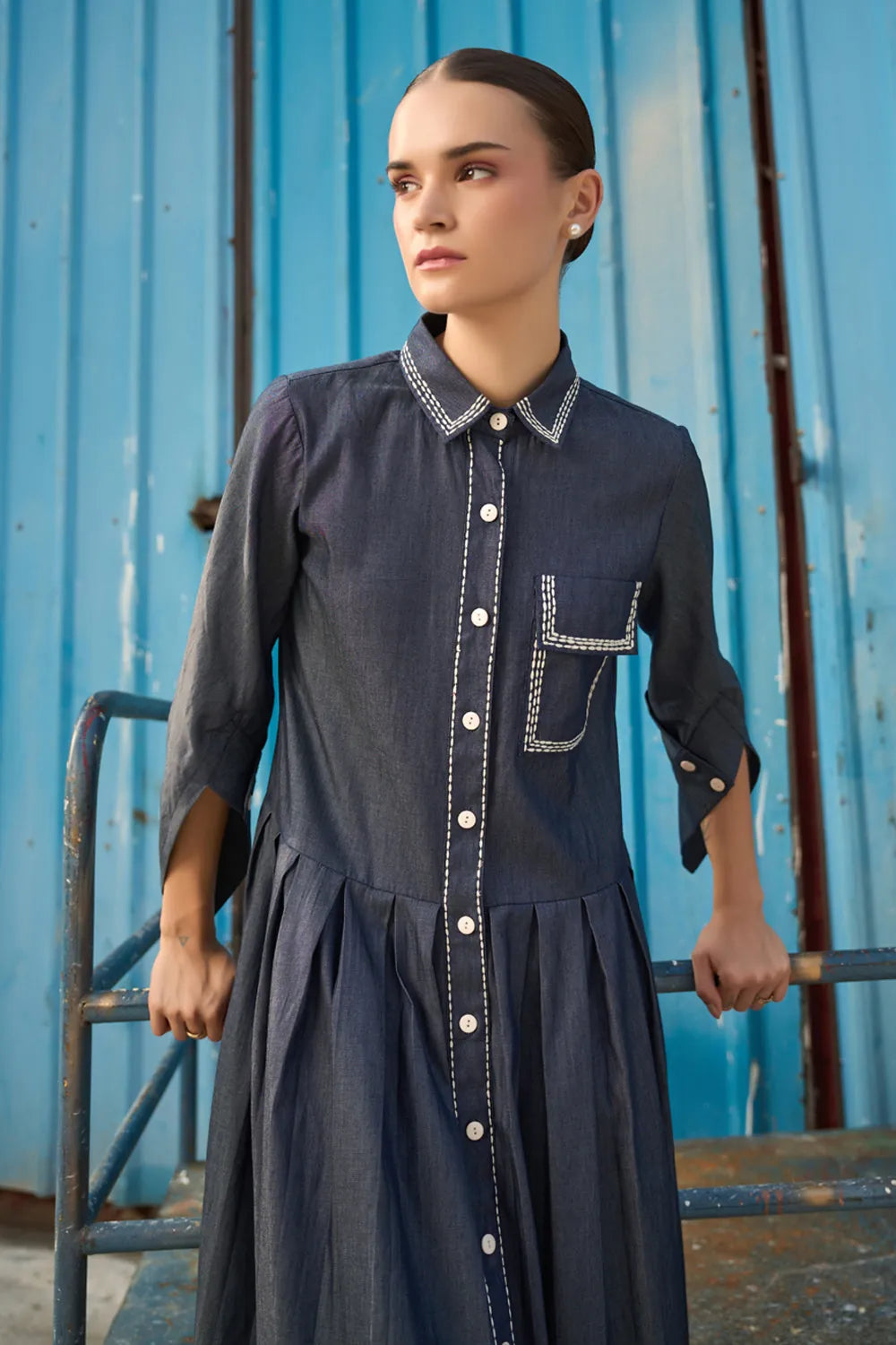 Closeup look of Elma Dress women's dark blue chambray midi dress featuring a structured shirt collar, button-down front, three-quarter sleeves with buttoned cuffs, a statement chest pocket with contrast stitching and a softly pleated skirt, styled for work meetings, casual outings and day-to-evening occasions.Woman wearing a dark blue dress with white buttons against a blue metal wall.