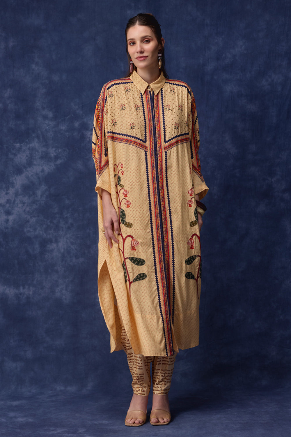A woman modeling a beige embroidered kurta with red, blue, and green floral details, paired with patterned pants, against a textured dark blue studio backdrop.