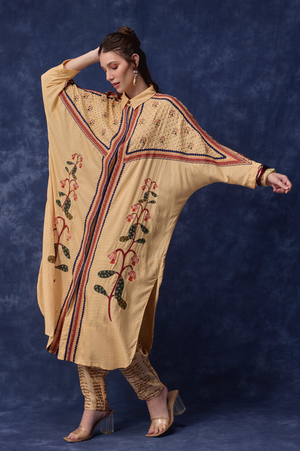 A woman modeling a beige kaftan-style tunic with red, blue, and green floral embroidery and striped border details, paired with patterned pants and clear-strap heels, against a dark blue textured backdrop.