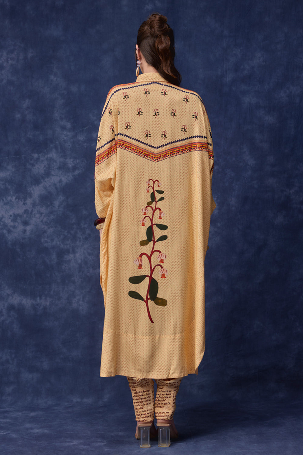 A woman viewed from behind wearing a long cream-colored embroidered tunic or kaftan with a floral vine motif down the back, standing against a mottled blue studio backdrop.