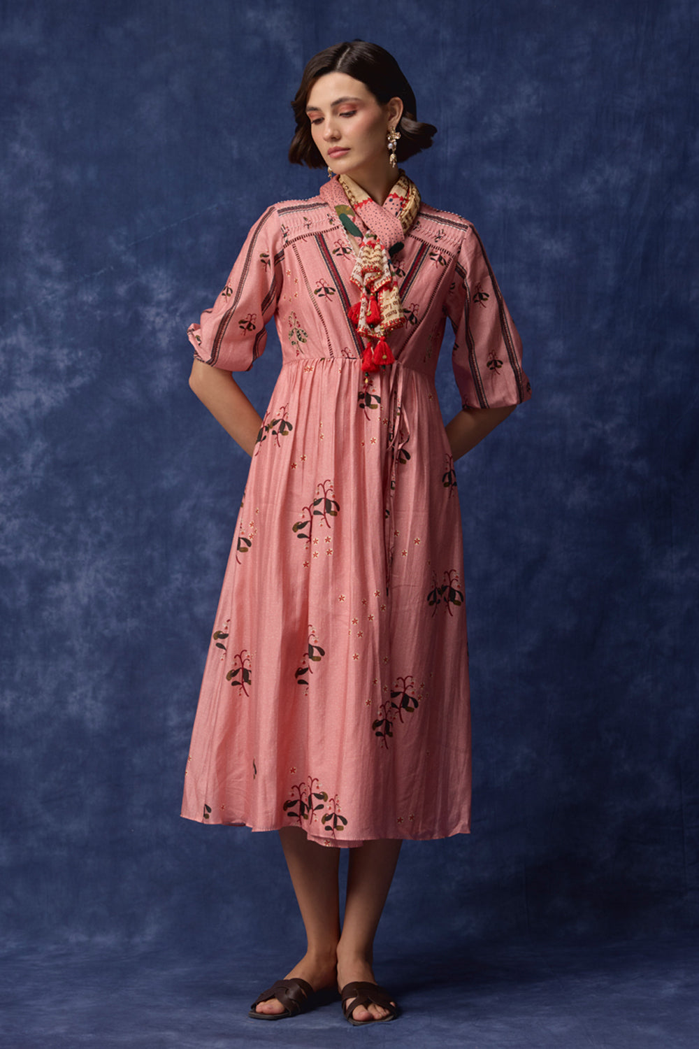 A woman modeling a pink midi dress with black floral embroidery, standing against a textured blue studio backdrop.