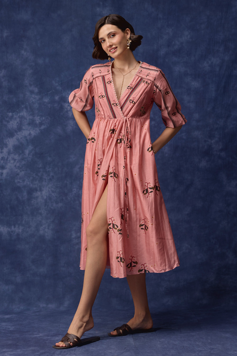 A woman models a pink midi dress with a cherry print, standing against a textured blue studio backdrop.