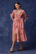 A woman models a pink midi dress with a cherry print, standing against a textured blue studio backdrop.