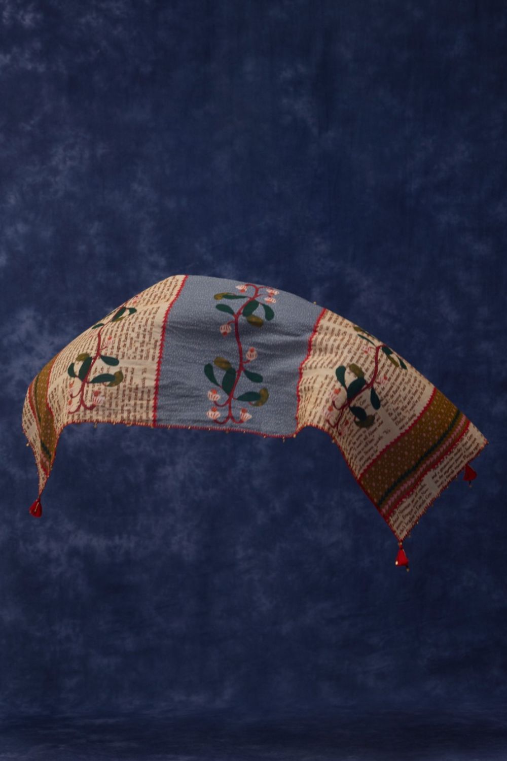 A handmade quilted fabric headband with a curved arch, featuring a central dusty blue panel with embroidered green leaves and pink flowers, flanked by cream and brown patterned borders, set against a mottled dark blue background.