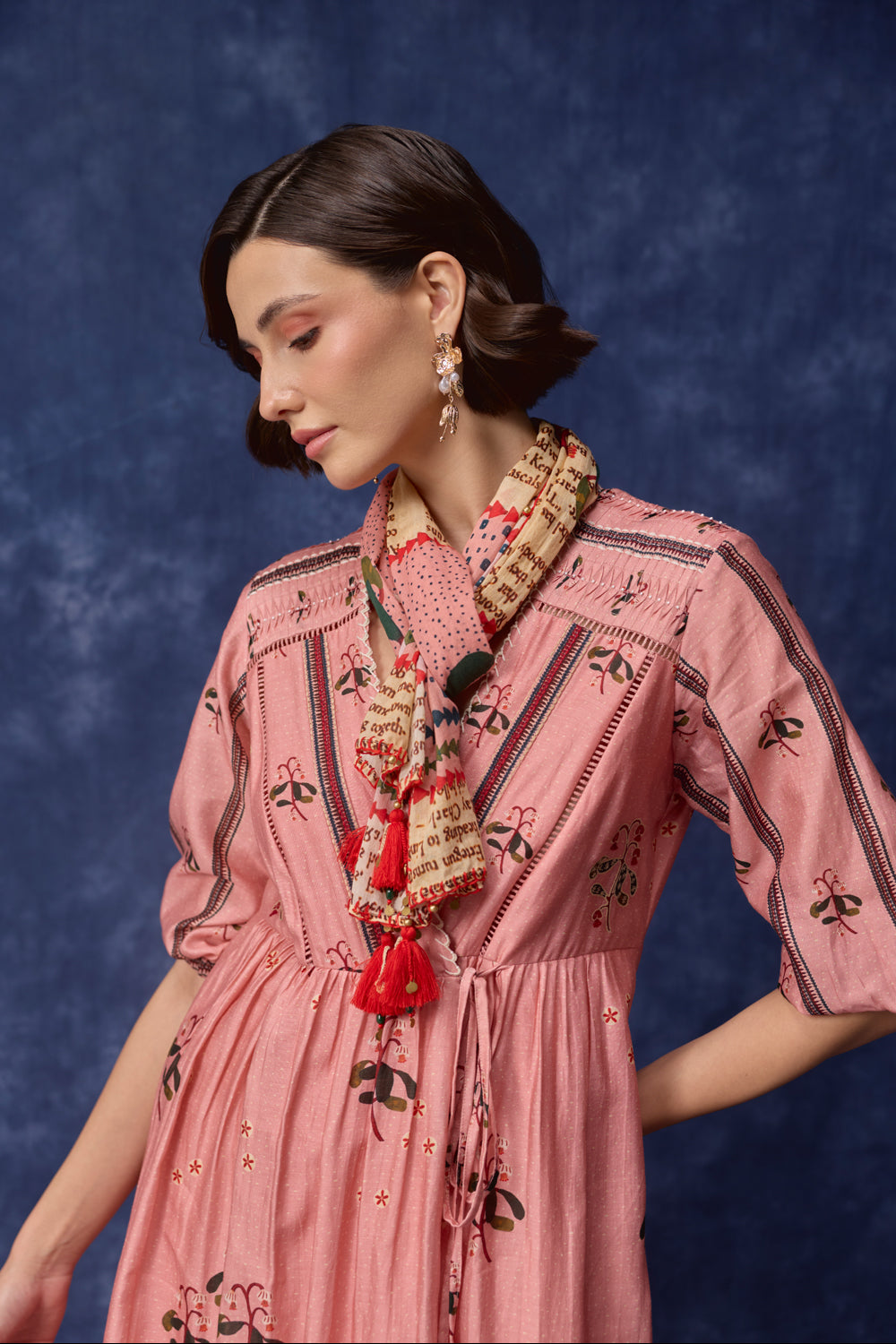 A woman wearing a pink embroidered dress with black floral and geometric patterns, styled with a patterned scarf and red tassel necklace, set against a dark blue textured backdrop.