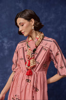A woman wearing a pink embroidered dress with black floral and geometric patterns, styled with a patterned scarf and red tassel necklace, set against a dark blue textured backdrop.