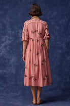 A woman stands with her back to the camera wearing a pink midi dress with black embroidered floral motifs and puff sleeves, set against a textured blue studio backdrop.