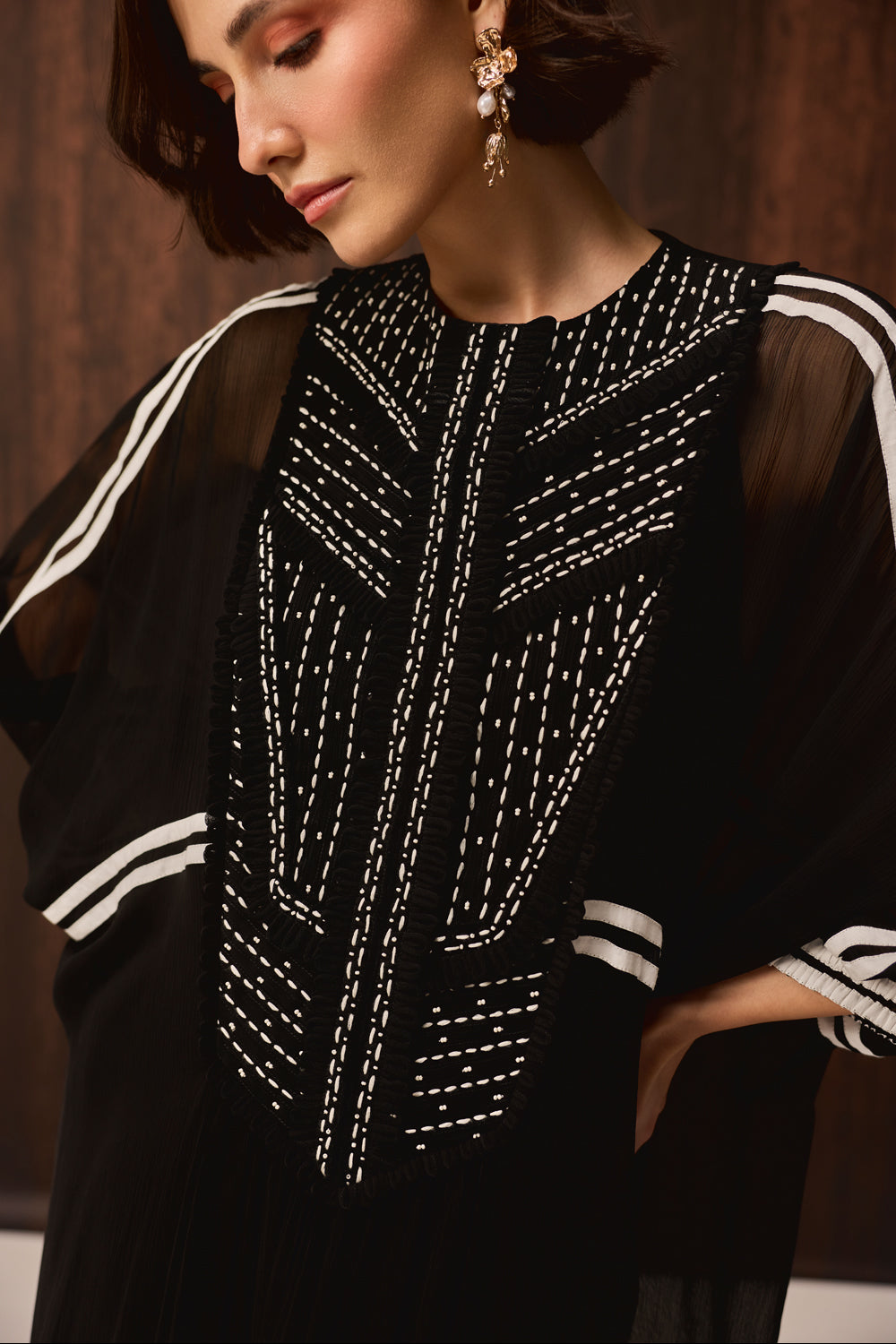 A close-up of a woman wearing a black embroidered tunic with white geometric beadwork and white stripe details on the sleeves, paired with dangling gold earrings.