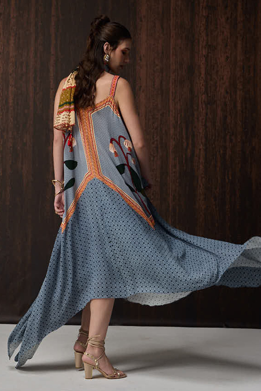 A woman modeling a flowing sleeveless dress with a dusty blue base, black polka dots, and orange embroidered details, shown from the back against a dark wood-paneled background.