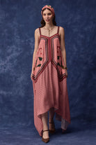 A woman modeling a pink sleeveless high-low dress with black polka dots, red and black embroidered details, and a matching pink headband, standing against a textured blue studio backdrop.