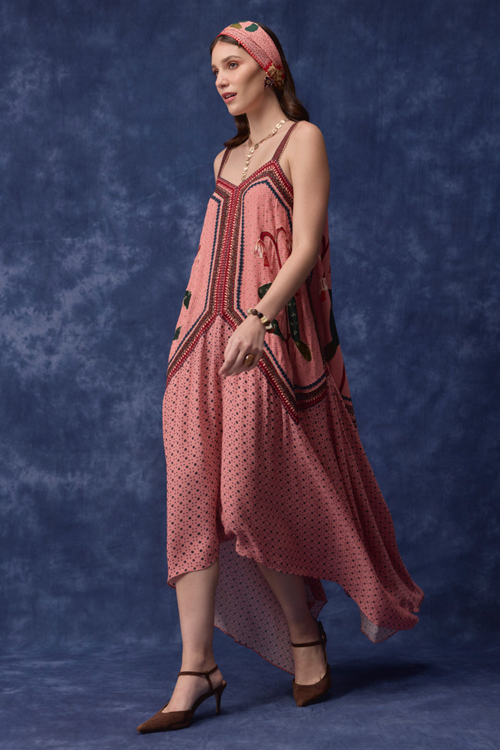 A woman models a pink sleeveless dress with a high-low hem, featuring a polka-dot pattern and embroidered details, paired with a matching pink headband against a textured blue studio backdrop.