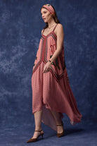 A woman models a pink sleeveless dress with a high-low hem, featuring a polka-dot pattern and embroidered details, paired with a matching pink headband against a textured blue studio backdrop.