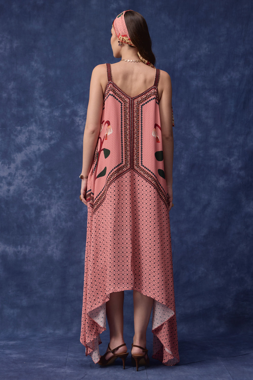 A woman stands with her back to the camera wearing a pink sleeveless high-low dress with dark brown geometric embroidery and a polka-dot pattern, paired with a matching pink headband, against a textured blue backdrop.