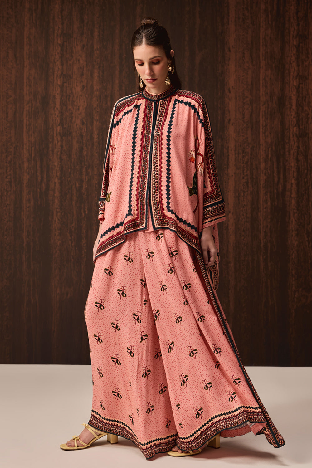 A woman models a traditional three-piece salwar kameez set in dusty pink, featuring black and gold border embroidery on the kameez and a matching printed salwar pant. She is standing against a dark brown wood-paneled background.