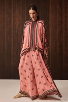 A woman models a traditional three-piece salwar kameez set in dusty pink, featuring black and gold border embroidery on the kameez and a matching printed salwar pant. She is standing against a dark brown wood-paneled background.