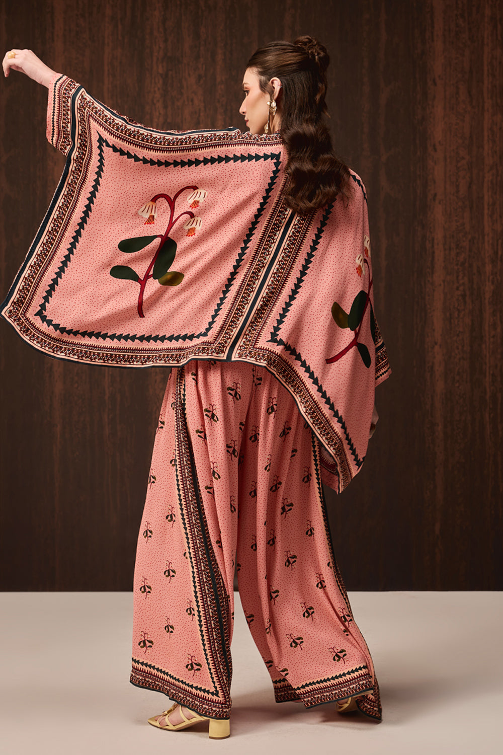 A woman modeling a pink traditional outfit with wide-leg pants and a matching embroidered shawl, posed against a dark wood-paneled background.