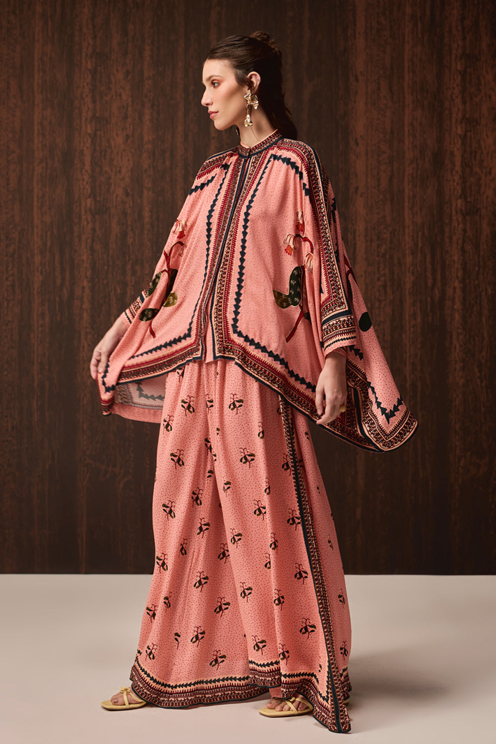 A model wearing a pink embroidered caftan with wide sleeves and a matching skirt, standing against a dark wood-paneled background.