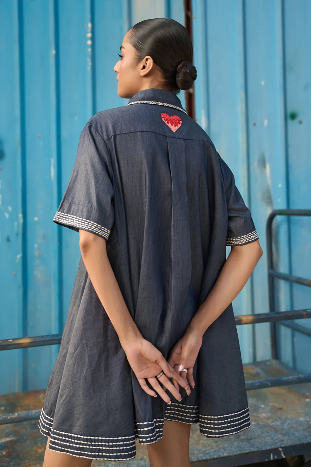 Back view of Rico Mini Dress women's dark blue chambray mini dress featuring a sharp pointed collar, short sleeves, a partial button-down front and contrast white stitching along the collar, sleeves, button placket and hem, finished with a loose flared A-line silhouette for casual outings, summer travel, relaxed daytime wear and casual dinners.