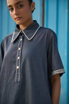 Closeup look of Rico Mini Dress women's dark blue chambray mini dress featuring a sharp pointed collar, short sleeves, a partial button-down front and contrast white stitching along the collar, sleeves, button placket and hem, finished with a loose flared A-line silhouette for casual outings, summer travel, relaxed daytime wear and casual dinners.