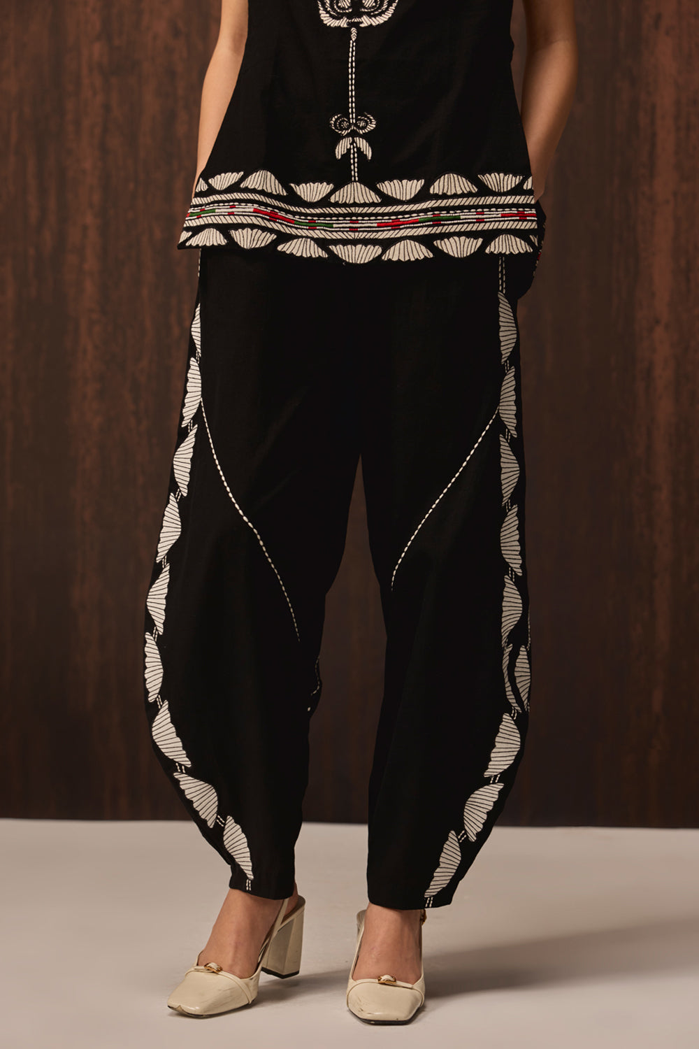 A person wearing black harem-style pants with white geometric embroidered patterns along the sides and hem, paired with a black sleeveless top, against a dark wood-paneled background.