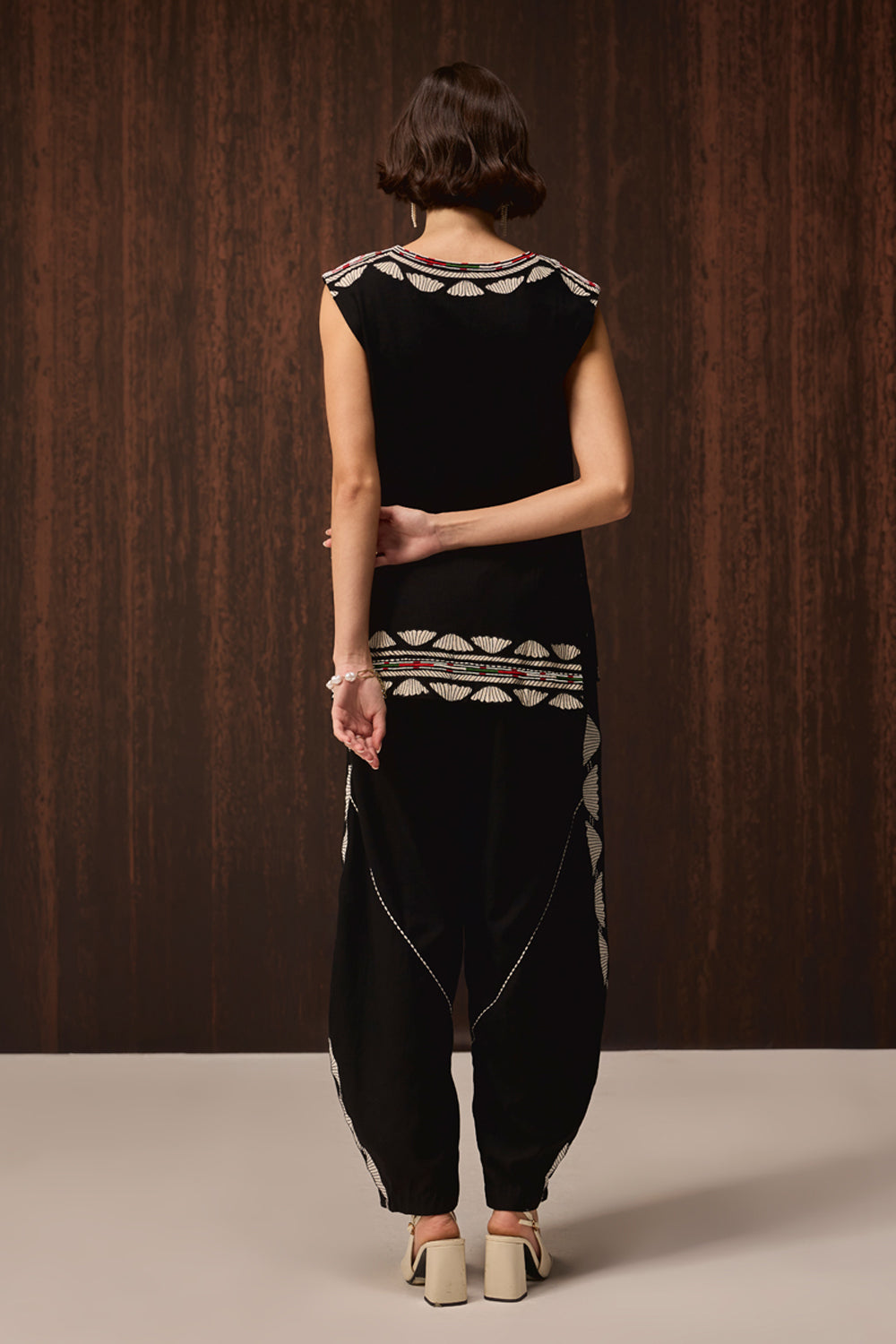 A model viewed from the back wearing a black sleeveless dress with cream and red geometric embroidery, paired with matching black harem-style pants and beige block-heel sandals, set against a dark wood-paneled background.