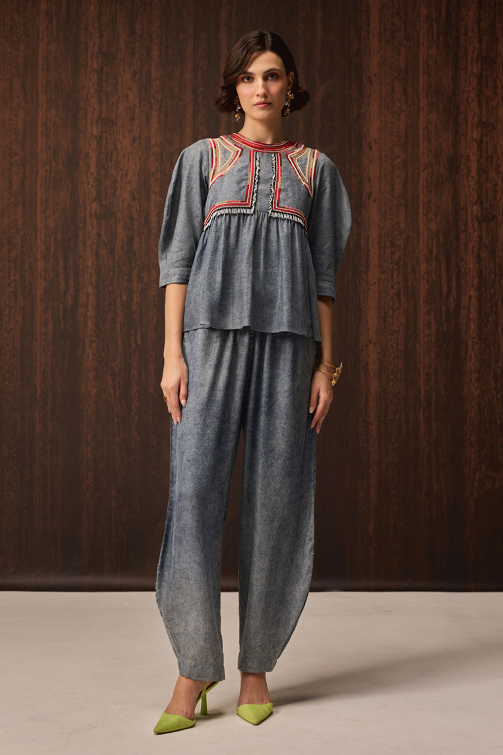 A woman models a matching two-piece outfit consisting of a slate blue-gray peplum top with red and yellow embroidery and matching wide-leg pants, set against a dark wood-paneled background.