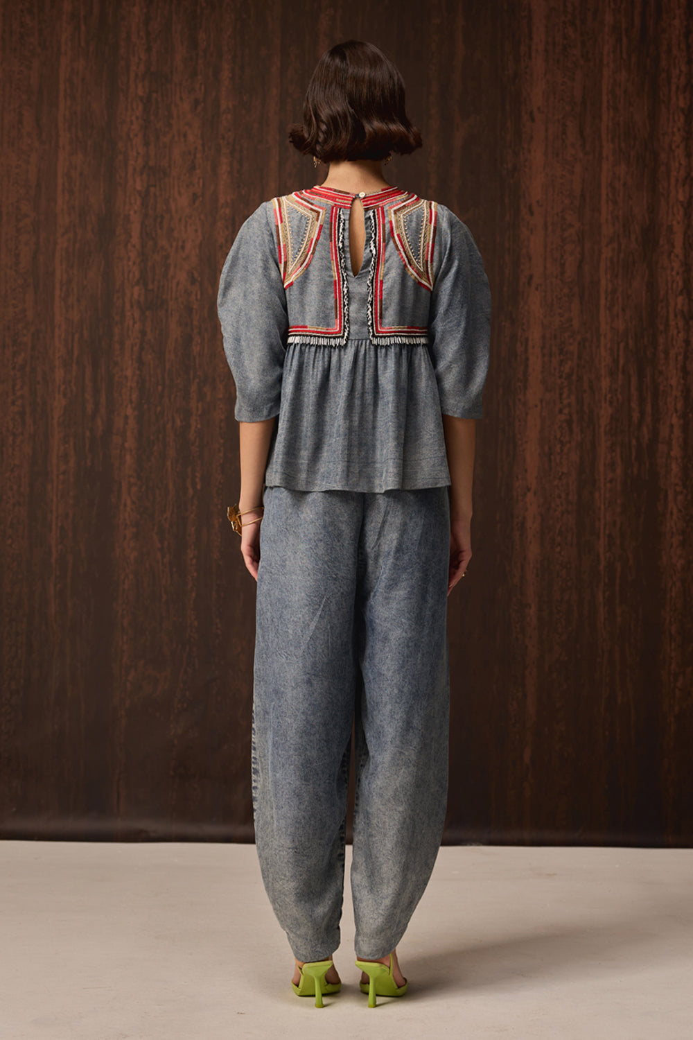 A model viewed from behind wearing a matching gray two-piece set consisting of a short-sleeve peplum top with colorful embroidered detailing on the back and wide-leg pants, set against a dark wood-paneled background.