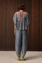 A model viewed from behind wearing a matching gray two-piece set consisting of a short-sleeve peplum top with colorful embroidered detailing on the back and wide-leg pants, set against a dark wood-paneled background.