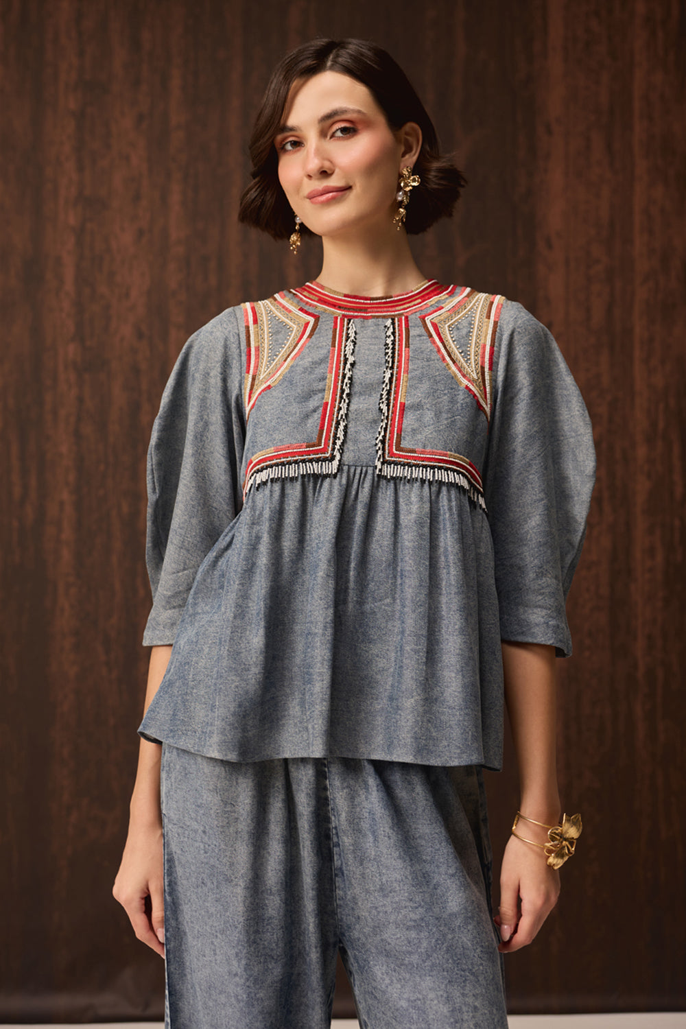 A woman modeling a blue-gray denim-like kurta set with red, yellow, and black embroidered detailing on the chest, paired with matching wide-leg pants, set against a dark wood-paneled background.