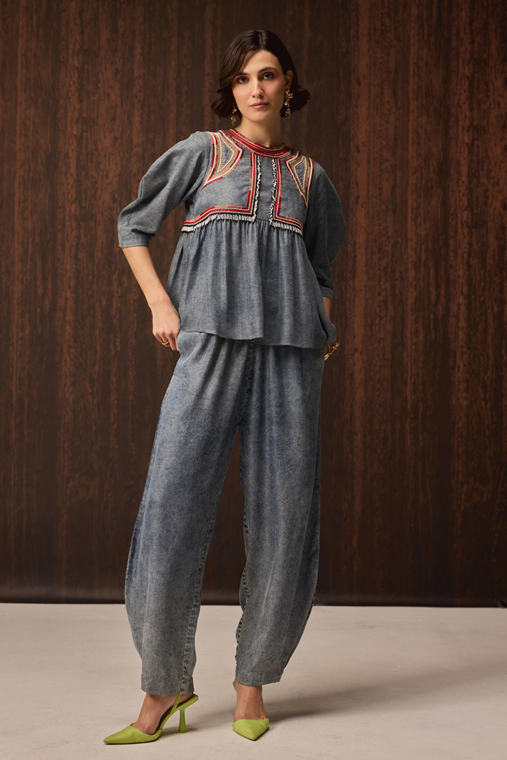 A woman models a two-piece denim set featuring a peplum top with red and yellow embroidered detailing at the neckline, paired with matching wide-leg pants, set against a dark wood-paneled background.