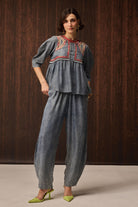 A woman models a two-piece denim set featuring a peplum top with red and yellow embroidered detailing at the neckline, paired with matching wide-leg pants, set against a dark wood-paneled background.