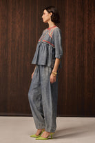 A woman stands in profile wearing a light blue denim two-piece outfit with embroidered detailing on the top and wide-leg pants, paired with lime green heels, against a dark wood-paneled background.