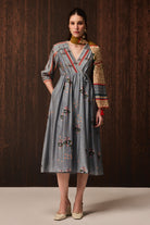 A woman models a gray midi dress with a floral print, featuring a shawl collar, tie waist, and a long patterned sleeve. She is posed against a dark wood-paneled background.