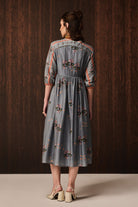 A woman viewed from the back wearing a gray midi dress with colorful floral embroidery and 3/4 sleeves, standing against a dark wood-paneled background.
