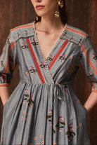 A woman wearing a gray wrap-style dress with colorful floral embroidery and red, orange, and green patterned trim, photographed against a warm brown background.