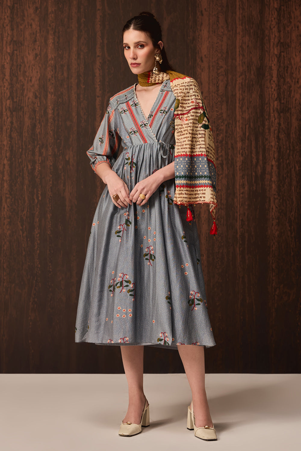 A woman models a gray midi dress with floral embroidery and a multicolor patterned shawl draped over her shoulder, set against a dark wood-paneled background.