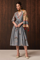 A woman models a gray midi dress with floral embroidery and a multicolor patterned shawl draped over her shoulder, set against a dark wood-paneled background.