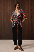 A model wearing a colorful patterned wrap-style shirt with tassel ties, black pants, and brown boat shoes, standing against a dark wood-paneled background.