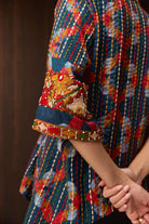 A close-up of a person wearing a colorful patterned tunic top with intricate embroidered detailing on the sleeve cuffs, set against a dark brown background.