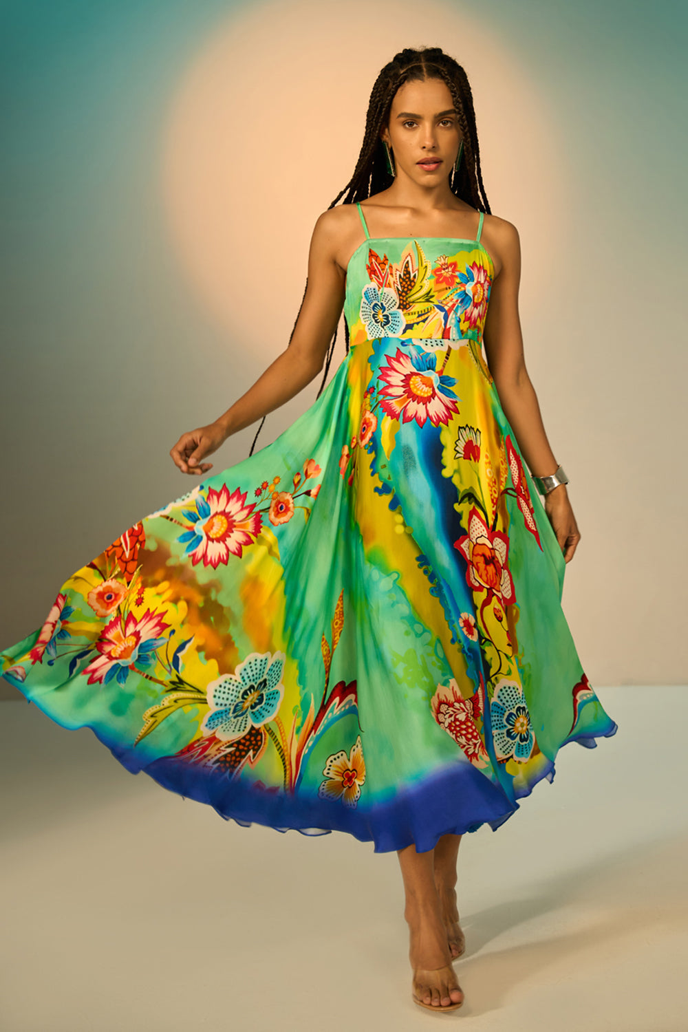 A woman modeling a vibrant, flowing midi dress with a colorful floral and abstract print. The dress features thin spaghetti straps and a ruffled hem, shown as she twirls against a studio backdrop with a warm spotlight.