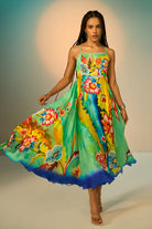 A woman modeling a vibrant, flowing midi dress with a colorful floral and abstract print. The dress features thin spaghetti straps and a ruffled hem, shown as she twirls against a studio backdrop with a warm spotlight.