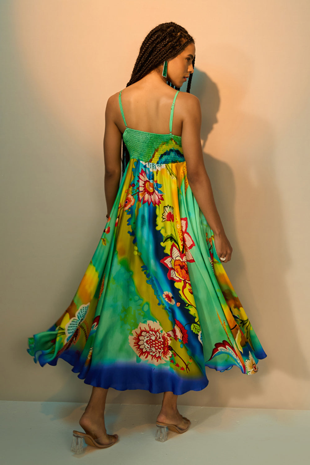 Back view of a woman modeling a vibrant, flowing midi dress with a colorful floral and abstract print. The dress features thin spaghetti straps and a ruffled hem, shown as she twirls against a studio backdrop with a warm spotlight.