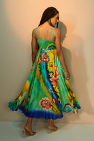 Back view of a woman modeling a vibrant, flowing midi dress with a colorful floral and abstract print. The dress features thin spaghetti straps and a ruffled hem, shown as she twirls against a studio backdrop with a warm spotlight.