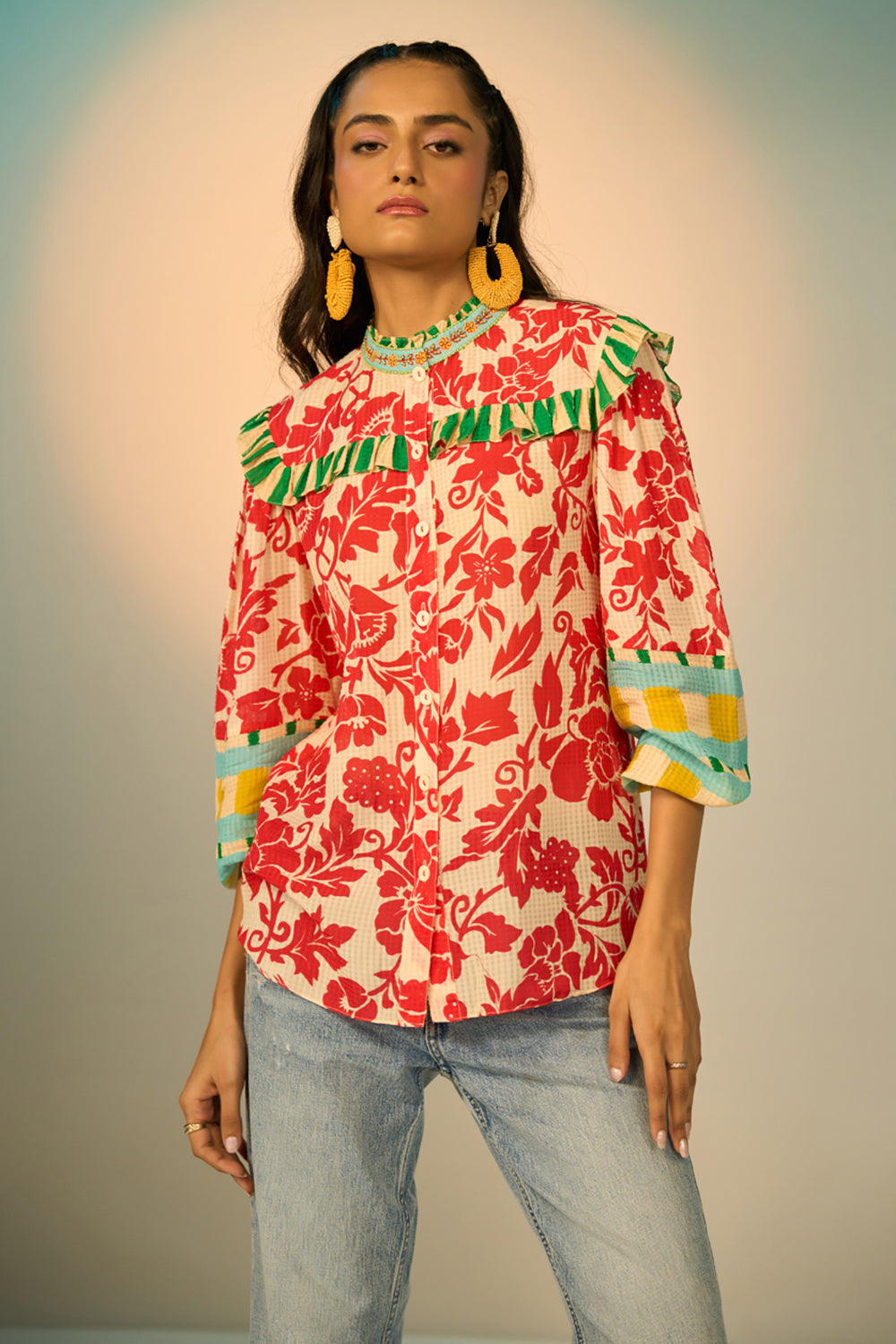 A woman modeling a vibrant red and cream floral print blouse with green and yellow accents, paired with light-wash jeans and gold hoop earrings, set against a soft beige studio background.