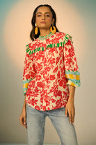A woman modeling a vibrant red and cream floral print blouse with green and yellow accents, paired with light-wash jeans and gold hoop earrings, set against a soft beige studio background.