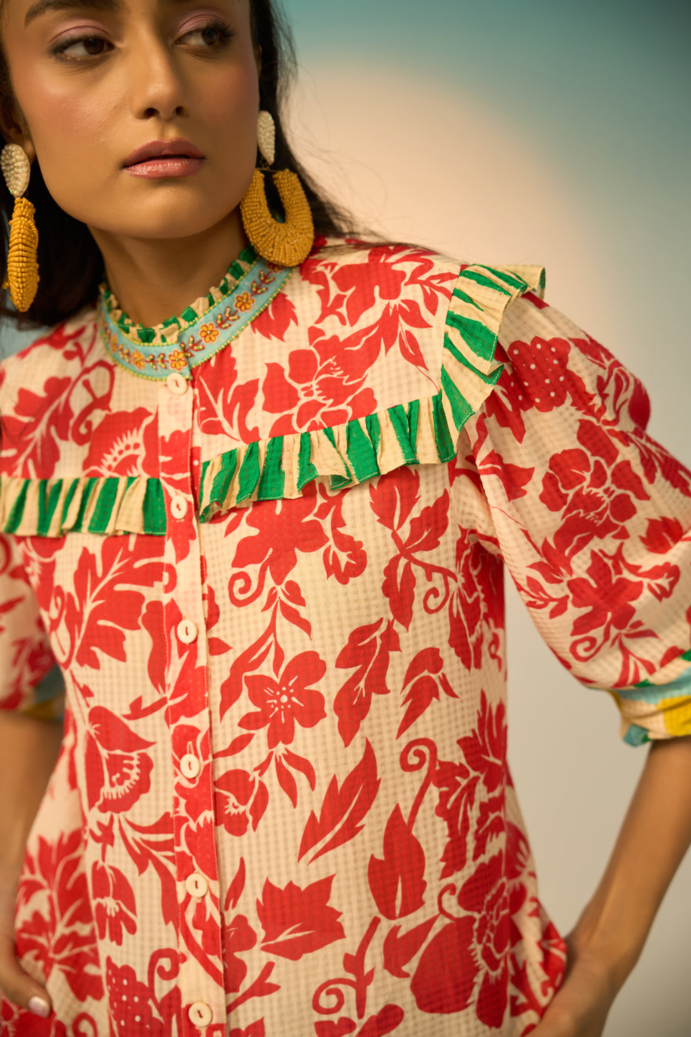 A woman modeling a vibrant red and cream floral print blouse with green and yellow accents, paired with light-wash jeans and gold hoop earrings, set against a soft beige studio background.