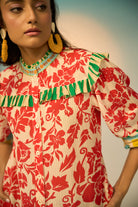A woman modeling a vibrant red and cream floral print blouse with green and yellow accents, paired with light-wash jeans and gold hoop earrings, set against a soft beige studio background.